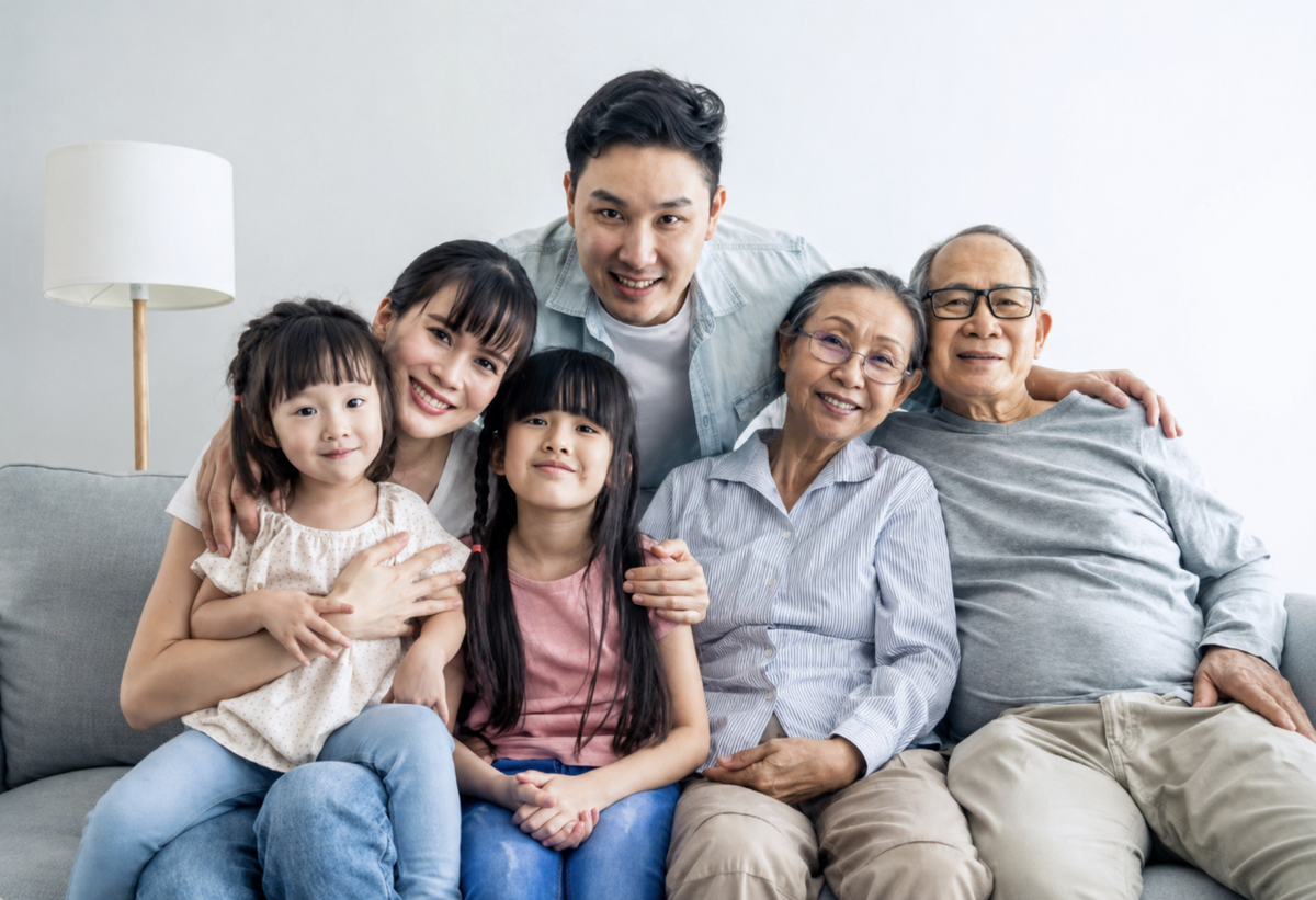 A family portrait of six people, including a young couple, two daughters, a grandfather, and a grandmother, sitting together on a gray sofa in a brightly lit living room.